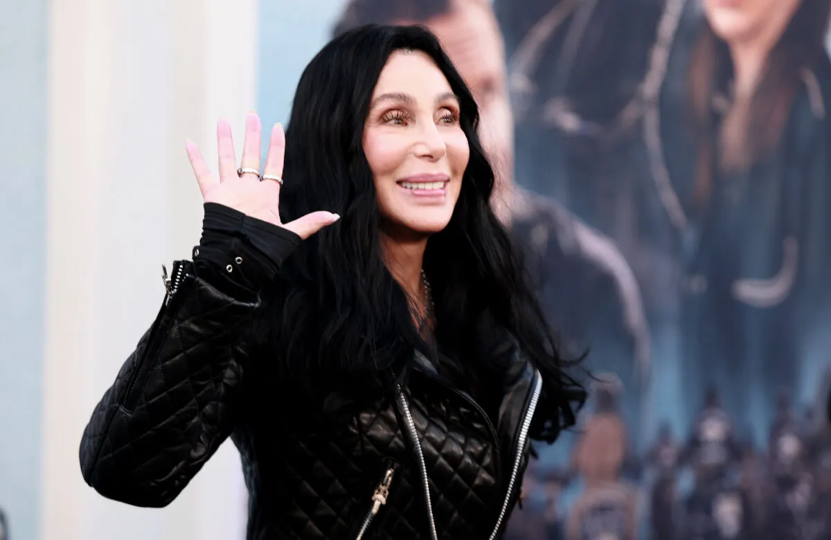 Cher wears a black jacket and waves.