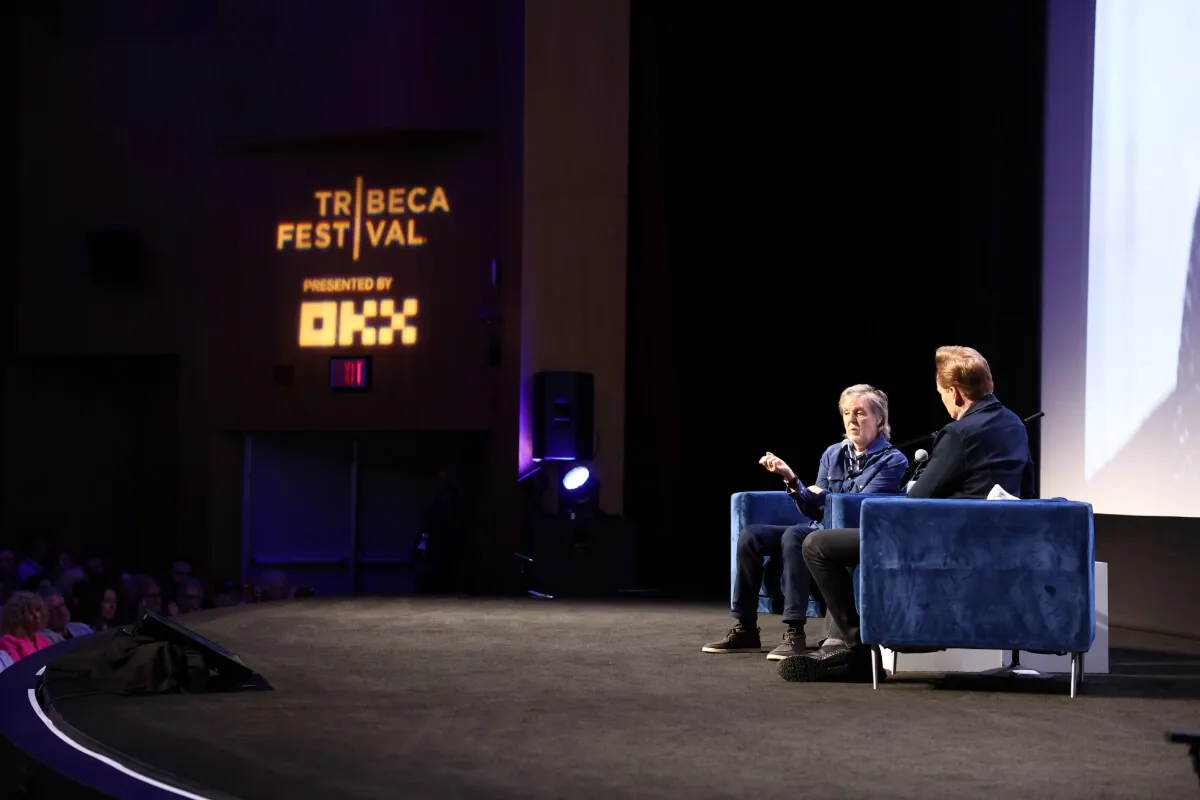 Paul McCartney and Conan O'Brien sit in armchairs on a stage together.