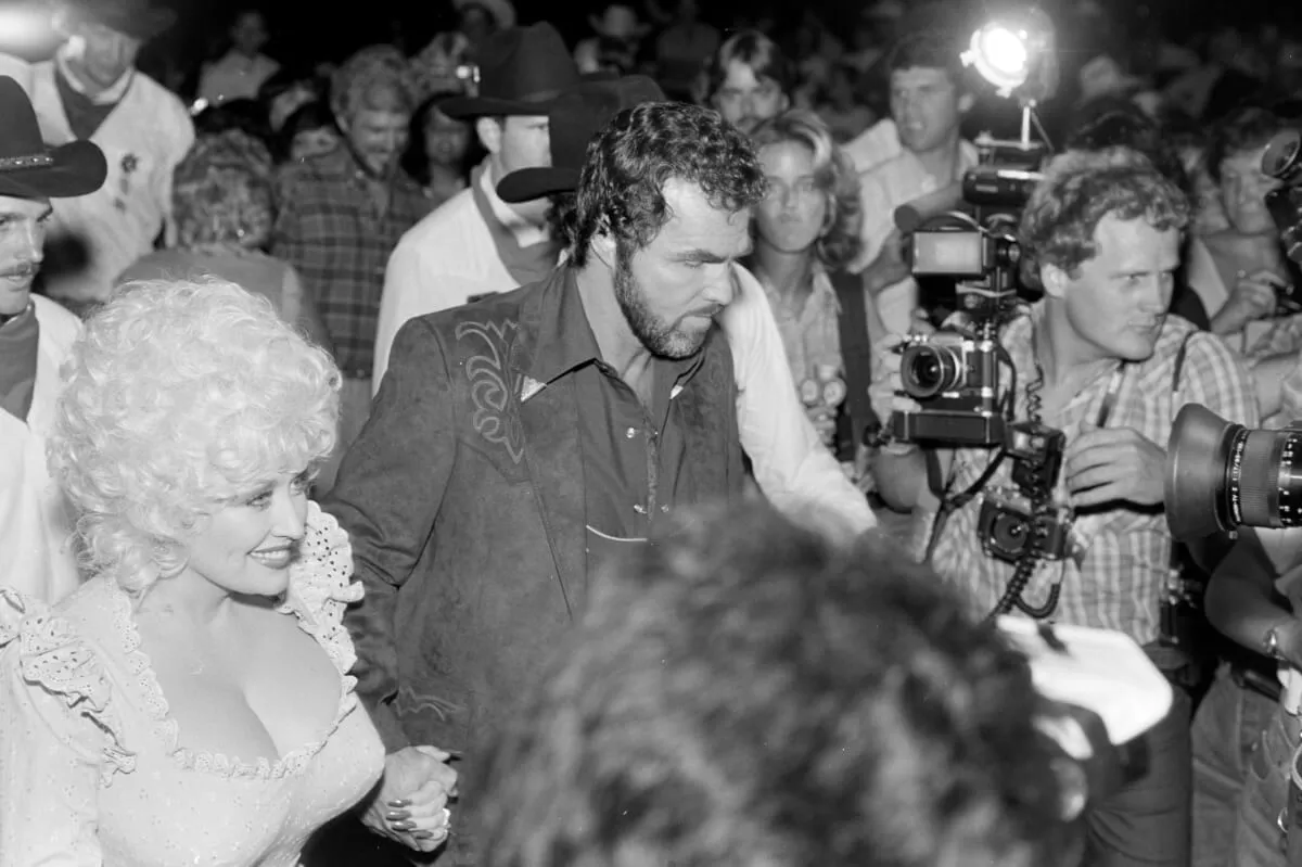 A black and white picture of Dolly Parton and Burt Reynolds holding hands while walking through a crowd.