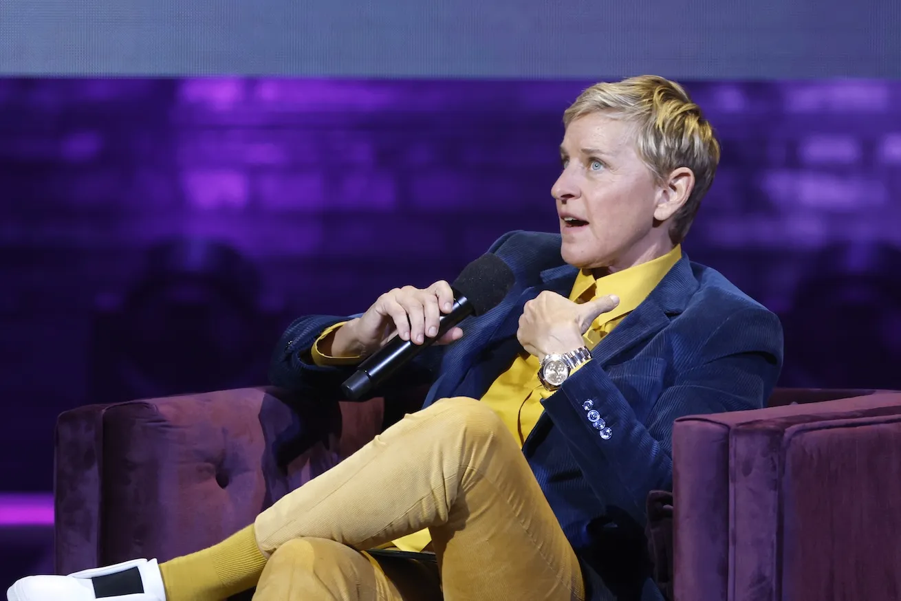 Ellen DeGeneres sitting in a chair with her legs crossed while speaking into a microphone