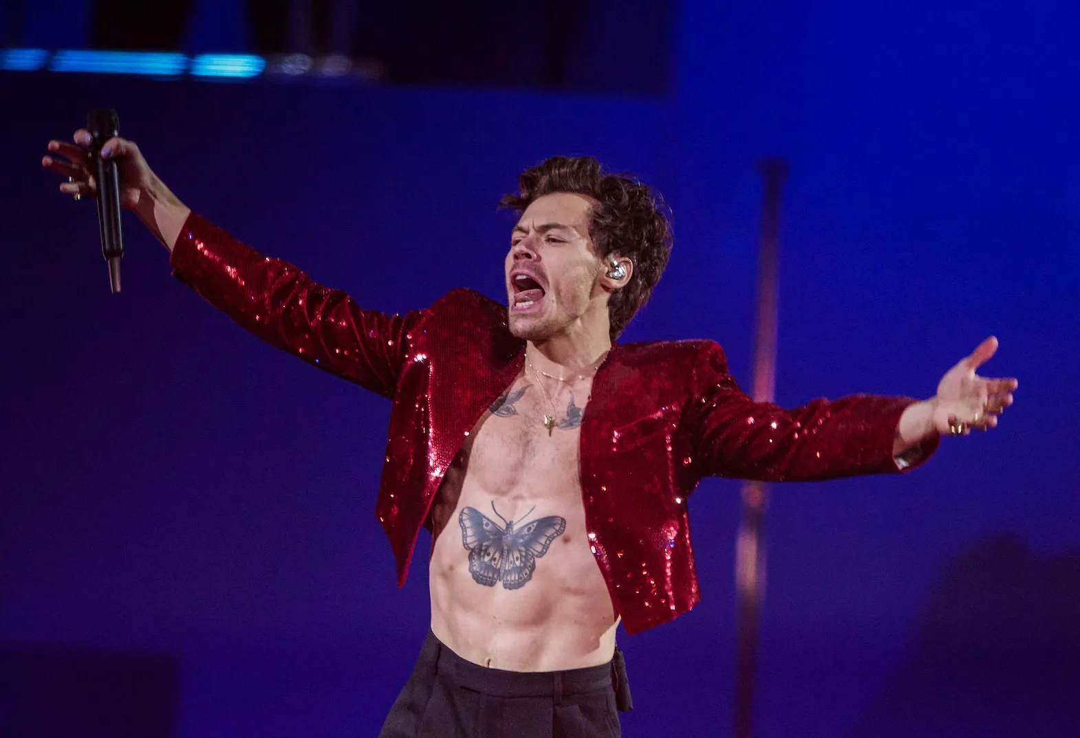 Harry Styles performs on stage shirtless while wearing a red jacket. He has his arms outstretched and his mouth open.