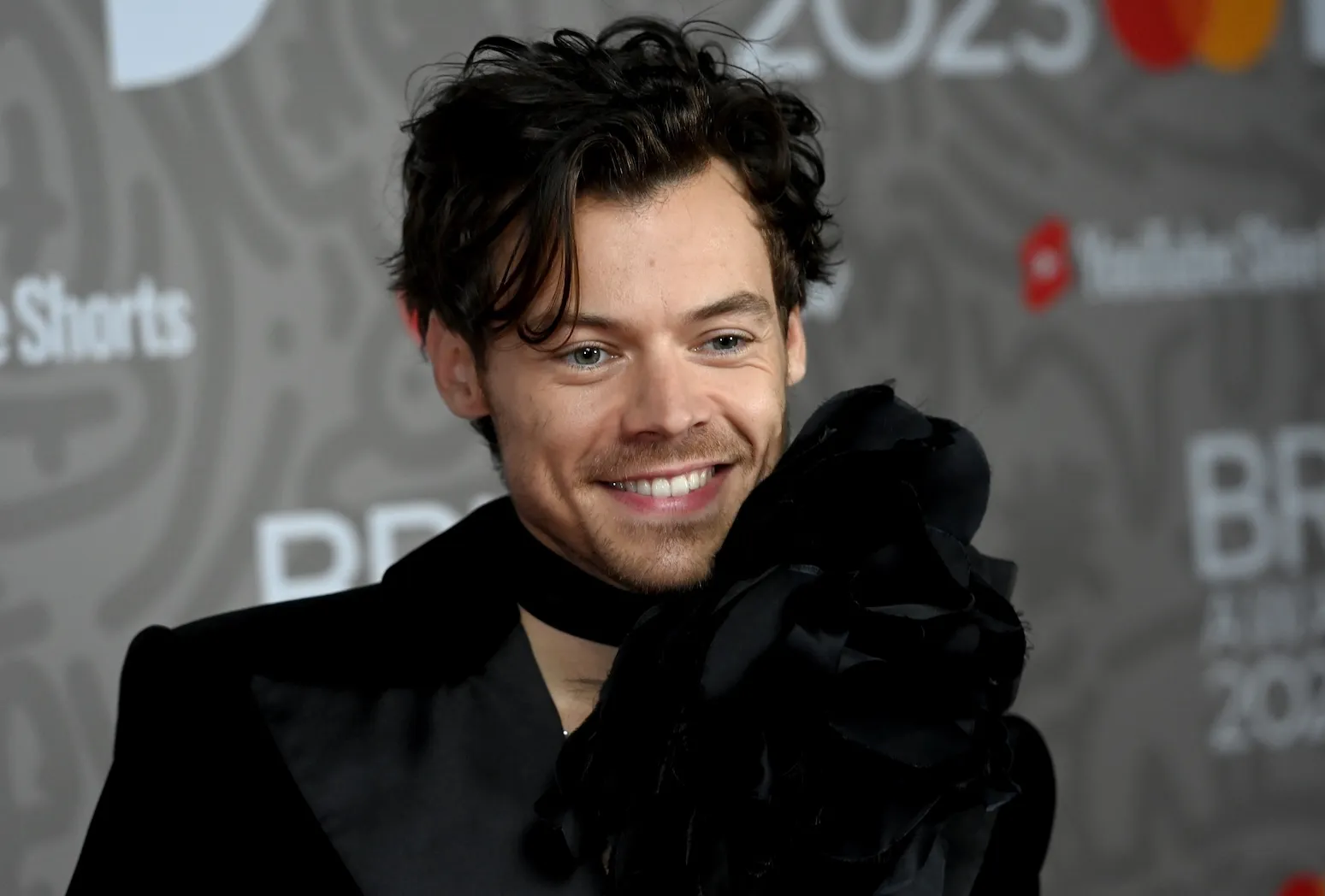 A close-up of Harry Styles smiling at the BRIT Awards in 2023. He's wearing a black coat and scarf.