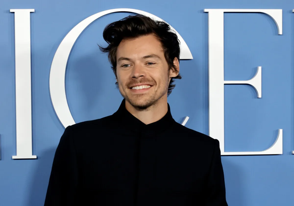 Harry Styles wears a black shirt and stands in front of a blue background.