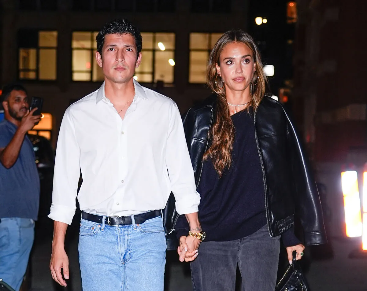 Danny Ramirez wears a white shirt and holds hands with Jessica Alba. She wears a black jacket.