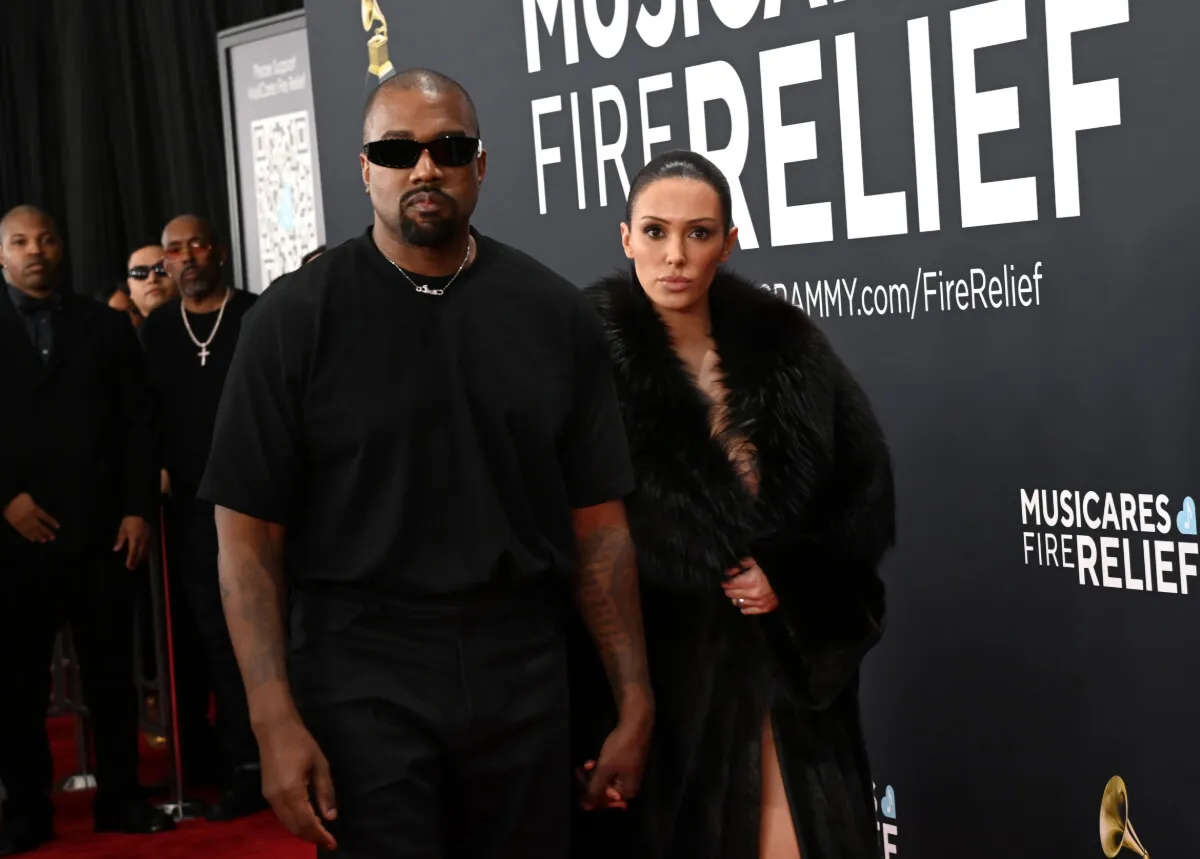 Kanye West wears a black shirt and sunglasses at the Grammys. Bianca Censori wears a black fur coat and stands behind him.