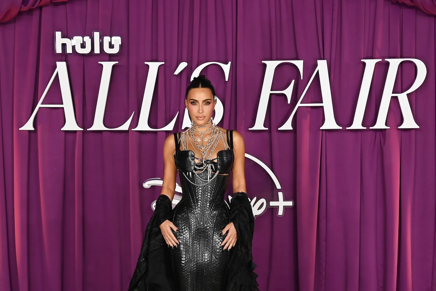Kim Kardashian in a black dress standing in front of a purple curtain with 'Hulu All's Fair' across it