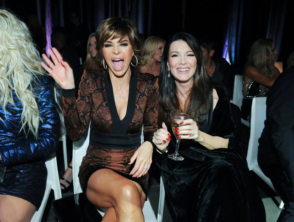 Lisa Rinna and Lisa Vanderpump sit together. Rinna wears a patterned dress and Vanderpump wears a black dress. She holds a glass of wine.