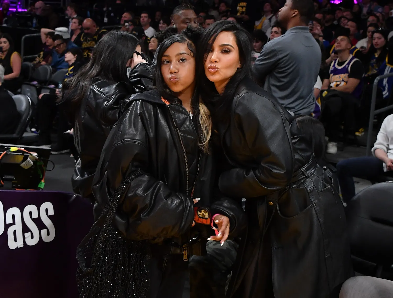 Kim Kardashian and North West posing and making kissing faces to the camera while at an NBA game. They're dressed in black and leaning in toward each other.