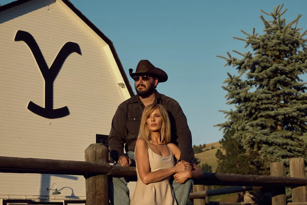 Rip, wearing a hat and sunglasses, standing behind Beth in front of the Yellowstone barn