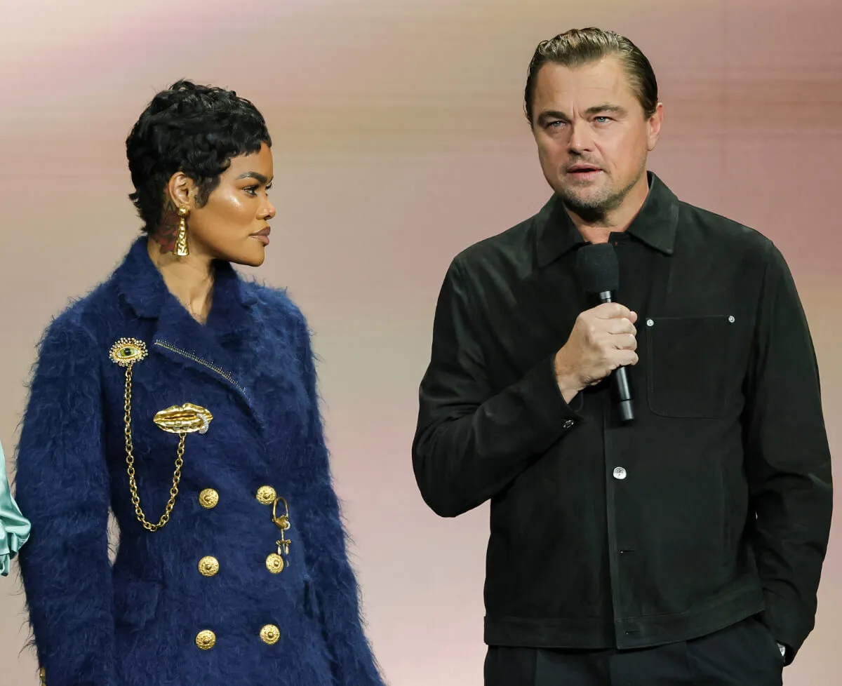 Teyana Taylor wears a blue coat and watches Leonardo DiCaprio speak into a microphone. He wears a black jacket.