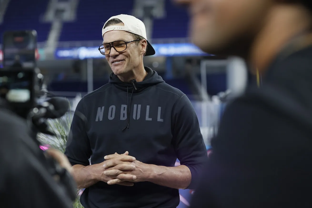 Tom Brady speaking with the media in January 2026. He has his hands clasped in front of him and he's wearing sunglasses and a baseball cap worn backwards.