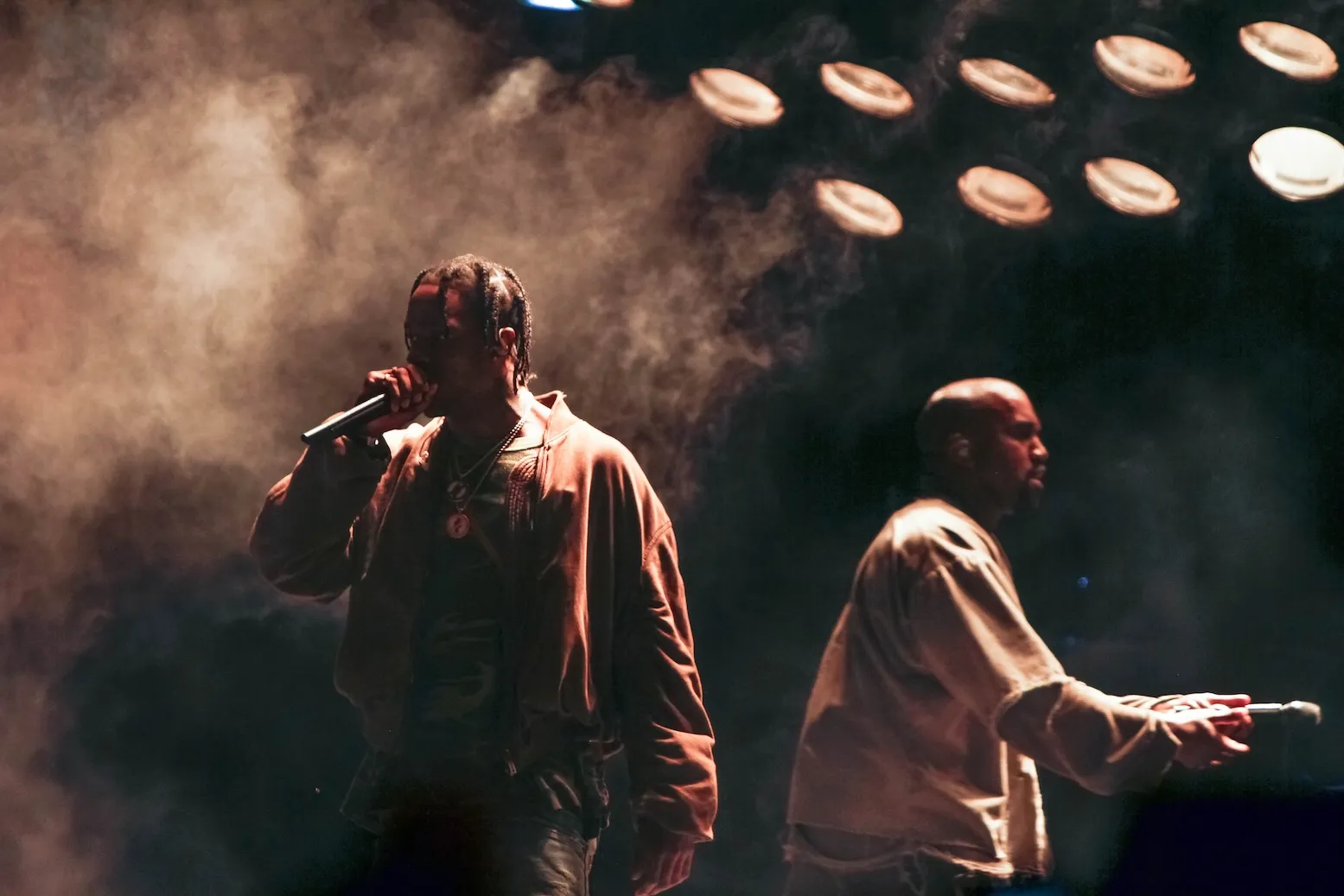 A dark and smoky photo of Travis Scott performing with Kanye 'Ye' West in the background onstage