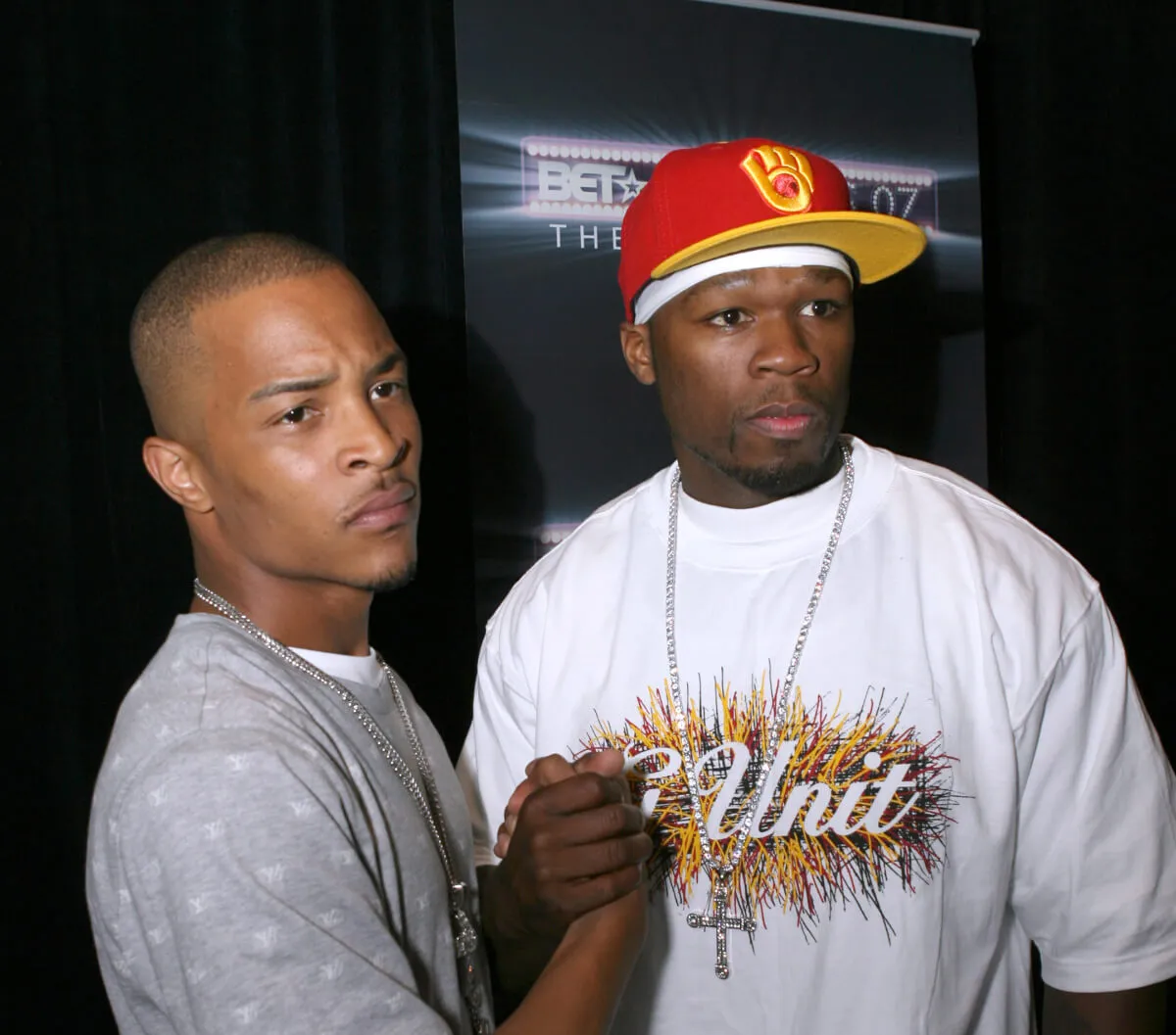 T.I. wears a gray shirt and shakes hand with 50 Cent. He wears a white shirt and red hat.