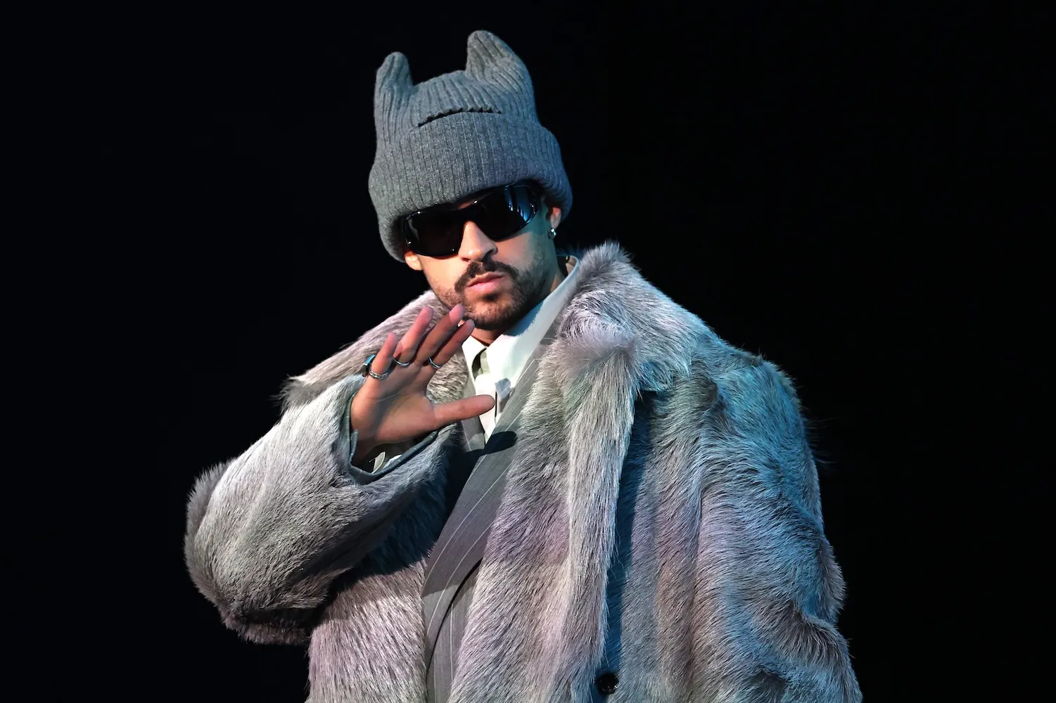 Bad Bunny with his hand up by his face against a black background. He's wearing sunglasses, a gray coat, and a gray hat.
