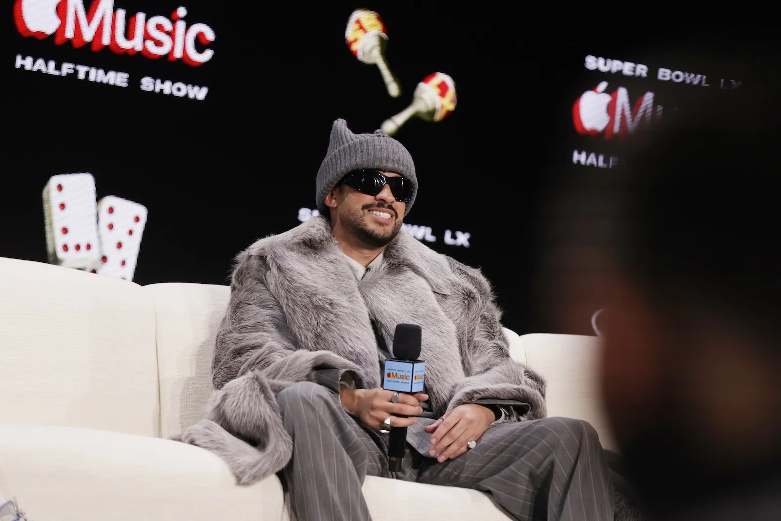Bad Bunny sitting on a white couch with a microphone in his hand. He's wearing a gray coat, a hat, and sunglasses.