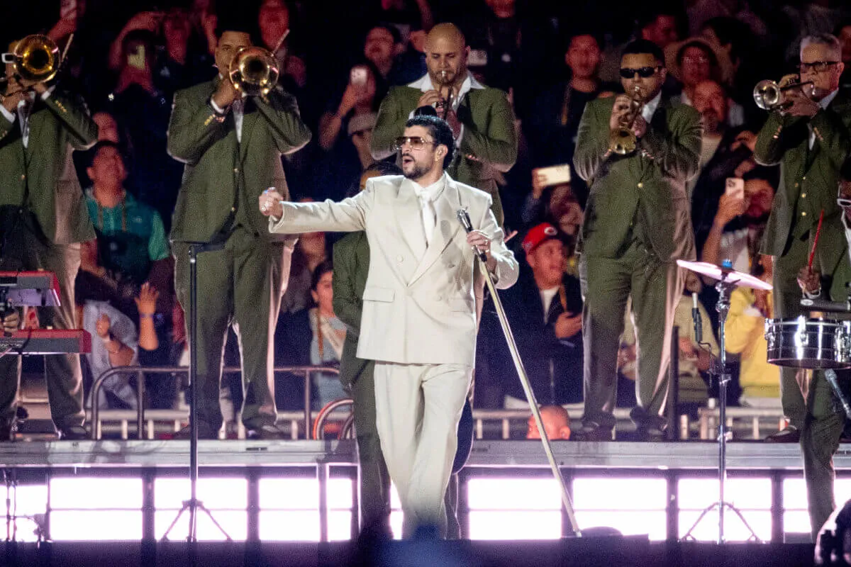Bad Bunny holds a microphone while onstage. He wears a tan suit. There is a brass band behind him.