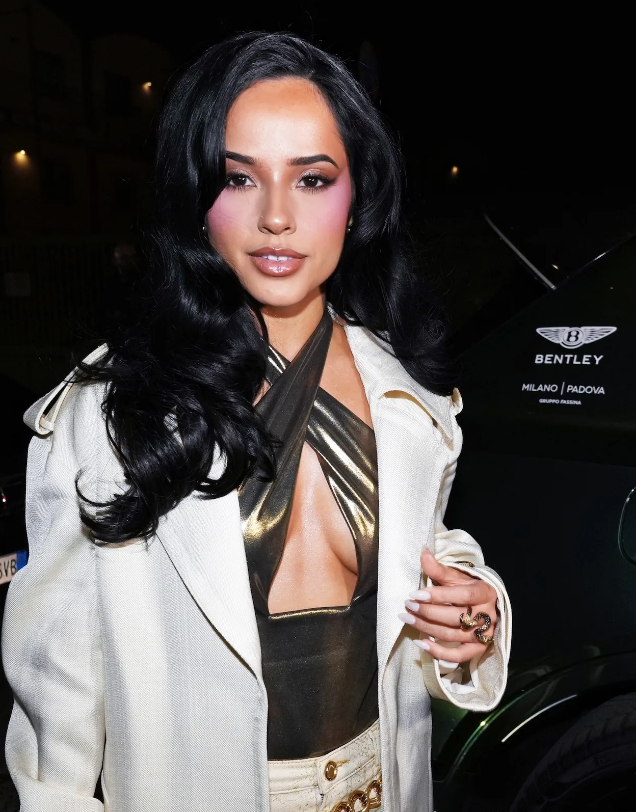 Becky G arrives in a Bentley to the Cavalli show during Milan Fashion Week 2026. She's wearing a cream-colored jacket over a metallic gold cross-front top.