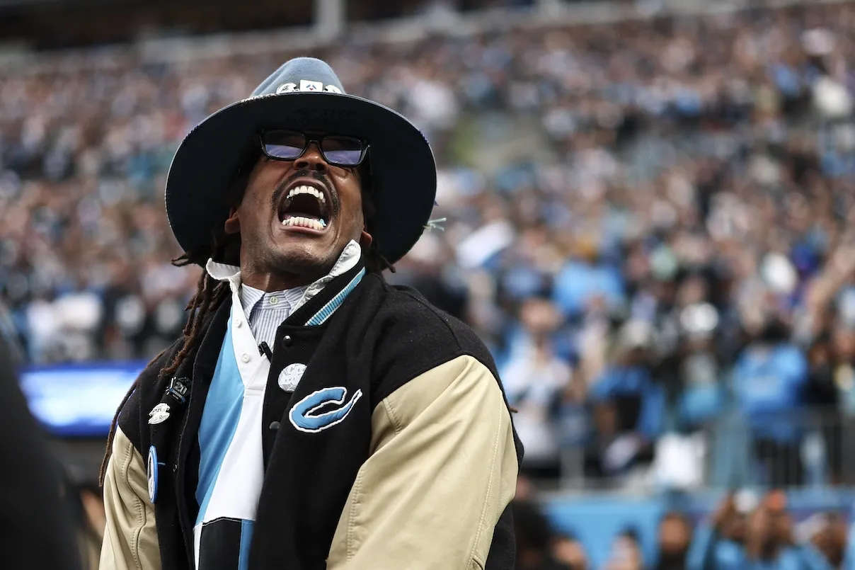Former Carolina Panthers quarterback Cam Newton screams during a Carolina Panthers and Los Angeles Rams game in January 2026. He's wearing a hat and sunglasses.