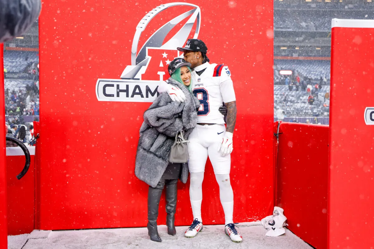 Stefon Diggs kisses Cardi B on the head at the AFC Championship game. Her hair is green and she wears a gray fur coat and baseball hat. He wears his football uniform.