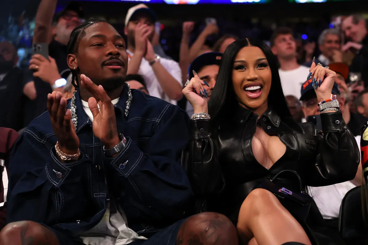 Cardi B and Stefon Diggs cheer at a basketball game. He wears a denim jacket and she wears a leather jacket.