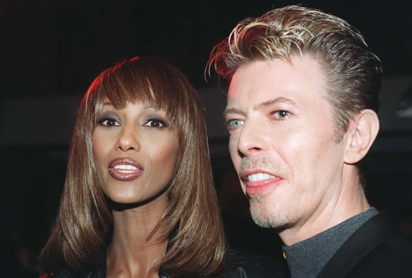 A close-up of David Bowie and his wife, Iman, in 1995. They are against a black background.