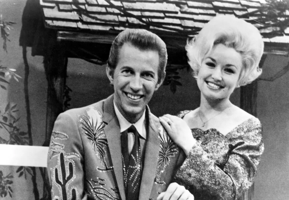 A black and white picture of Porter Wagoner and Dolly Parton posing together in front of a house set.
