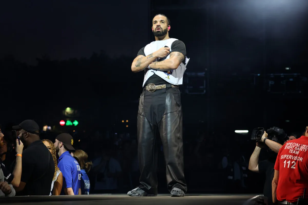 Drake stands on a stage with his arms crossed. He holds a microphone.