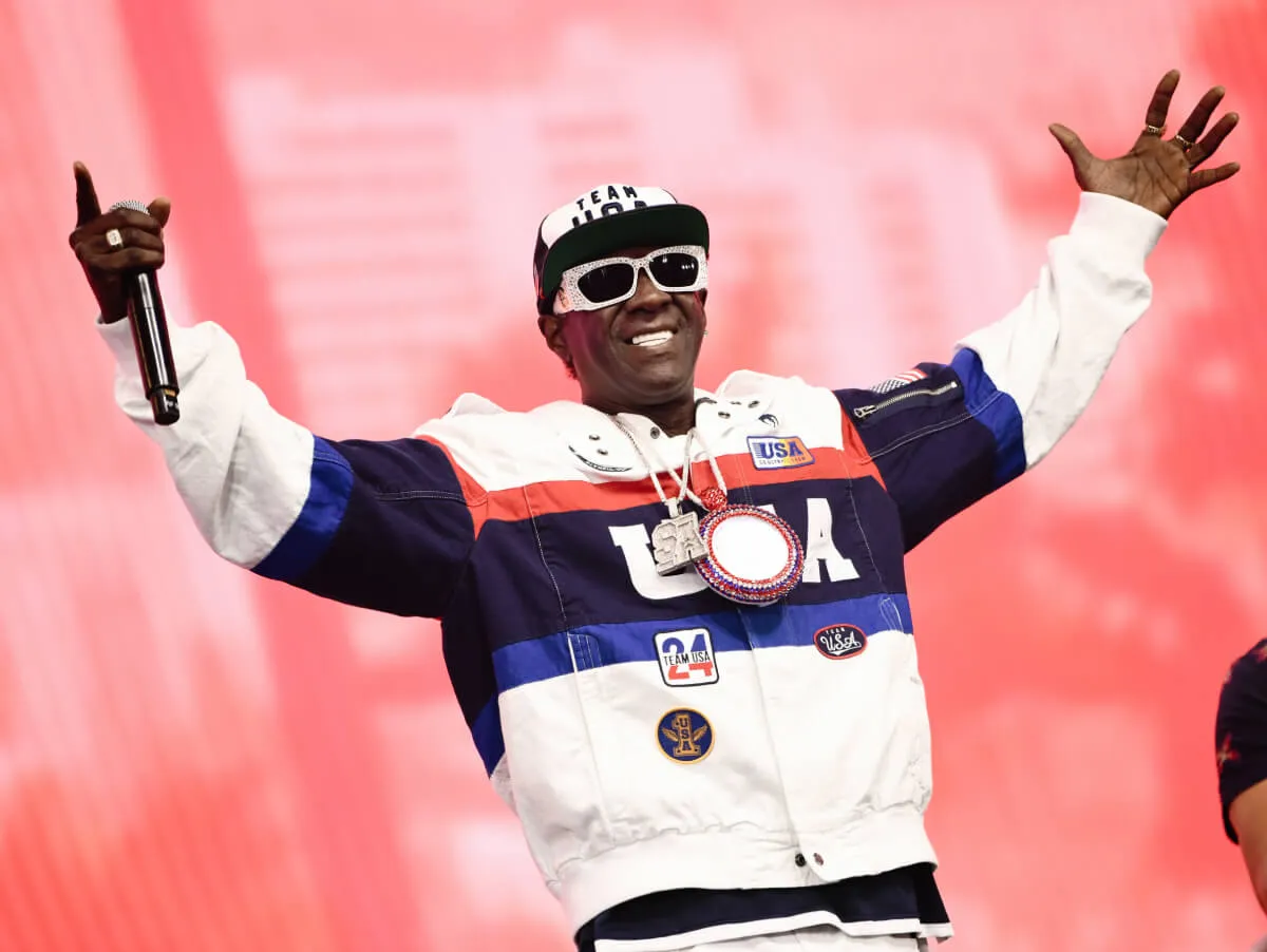 Flavor Flav wears a USA jacket, sunglasses, and a hat. He holds up a microphone.