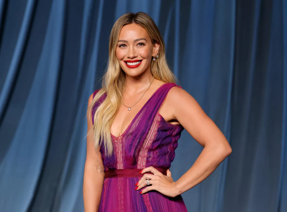 Hilary Duff wears a purple dress and stands with her hand on her hip.
