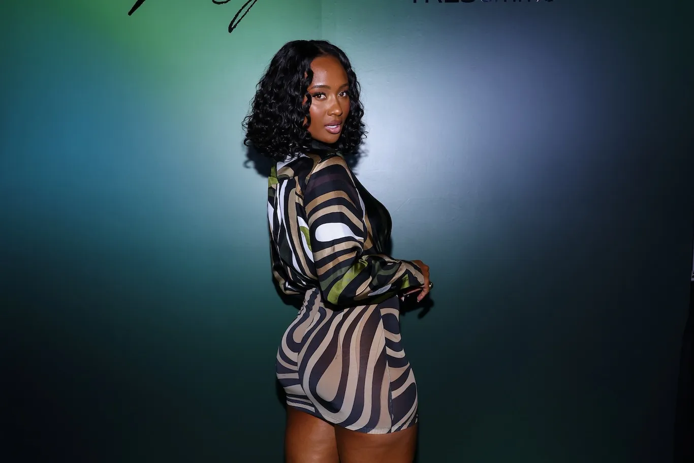 Kayla Nicole at an event in September 2025. She's wearing a short, long-sleeve striped dress and is looking over her shoulder to the camera.