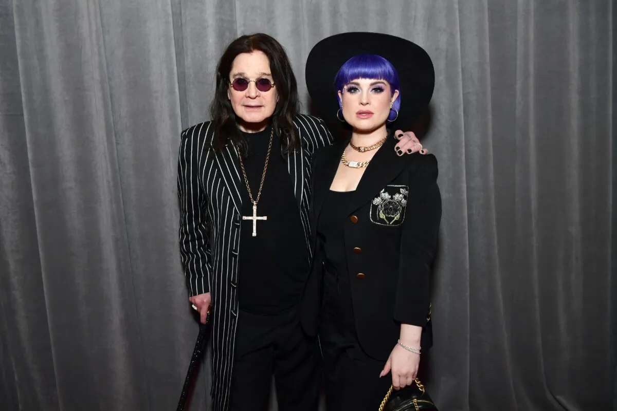 Ozzy Osbourne wears a pinstriped suit and sunglasses. He stands with his arm around Kelly Osbourne's shoulders. She wears a black outfit and hat. Her hair is blue.O