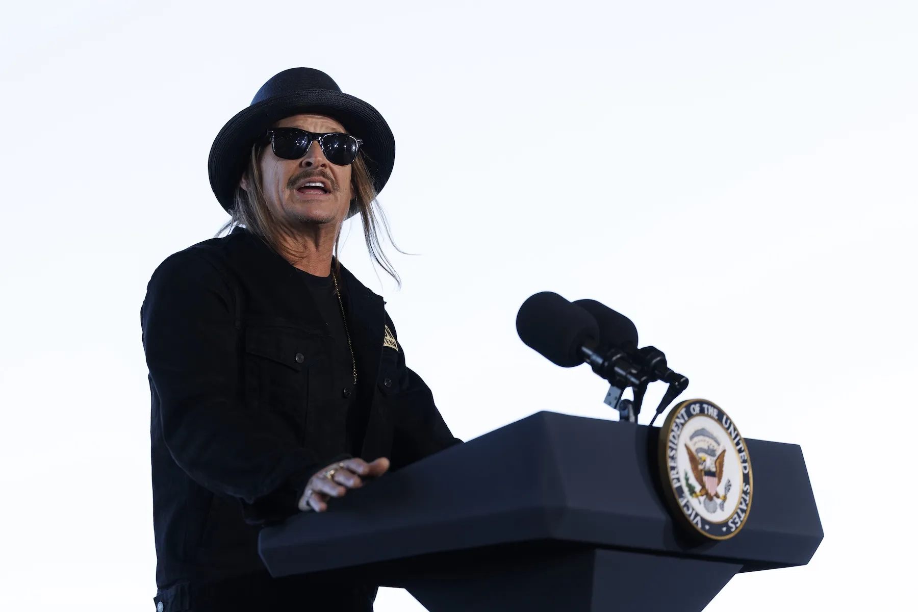 Musician Kid Rock standing at a podium. He's wearing a black hat and sunglasses.