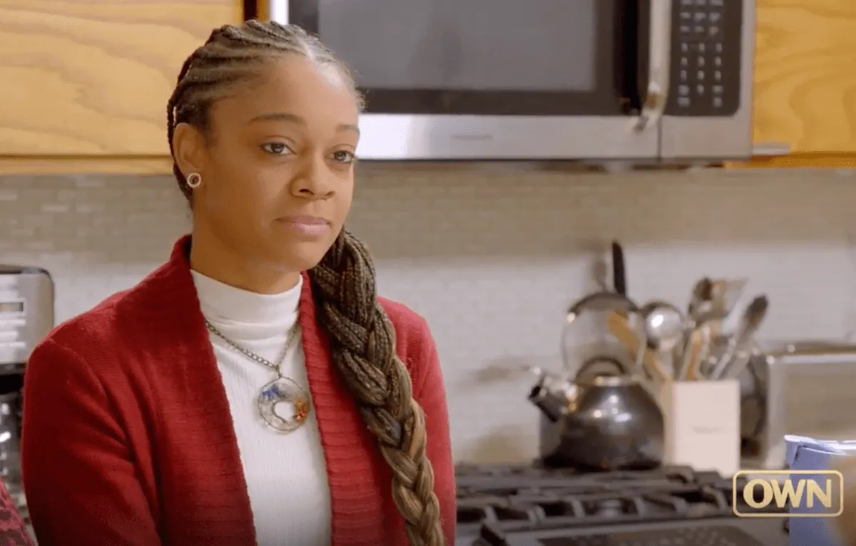 Nyeela, with her hair in braids and wearing a red jacket and white turtlneck, sitting at a kitchen table in OWN's 'Maxxed Out'