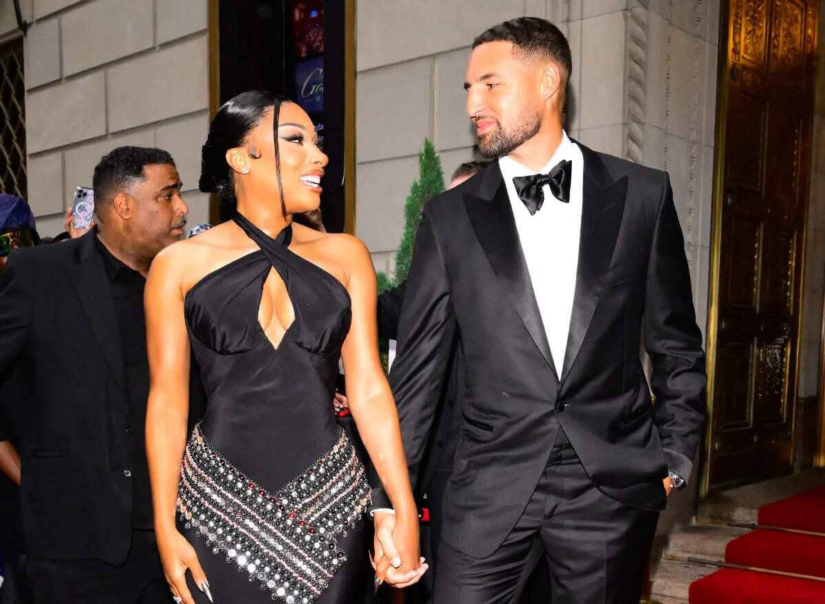 Megan Thee Stallion wears a black dress and holds hands with Klay Thompson. He wears a tuxedo.