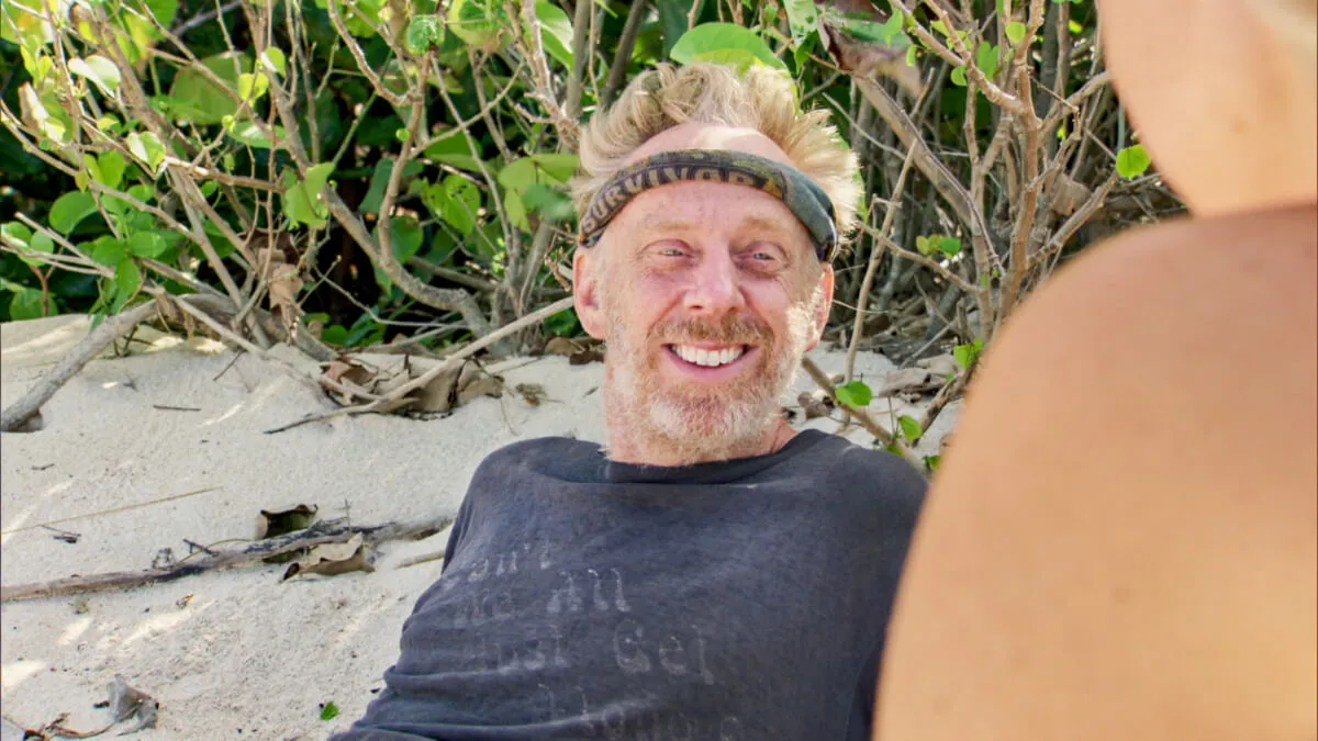 Mike White sits on a beach on 'Survivor.' He wears a gray shirt and has a buff around his head.