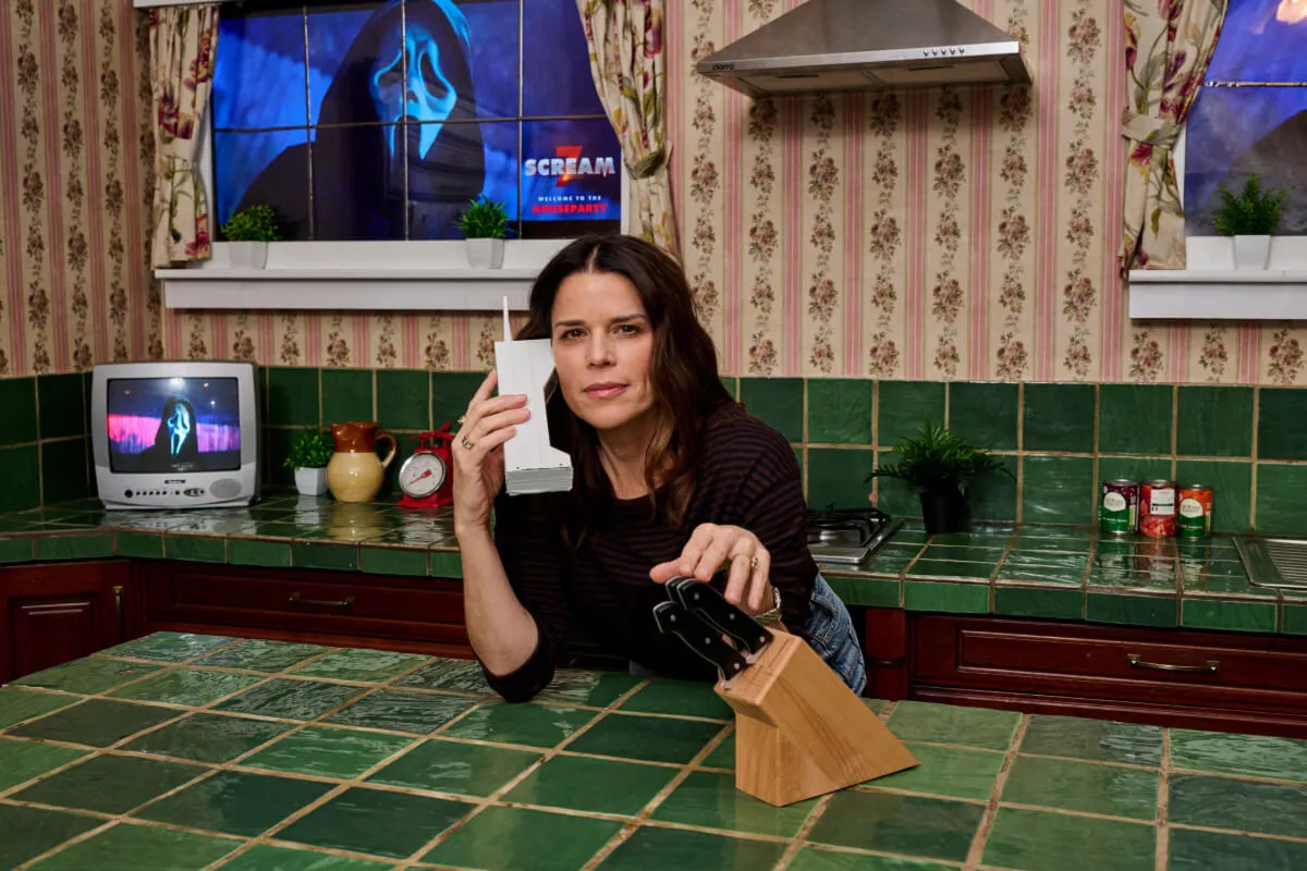 Neve Campbell leans over the kitchen counter in the 'Scream' house. She speaks into the phone and touches a block of knives. Ghost Face is in the window behind her.