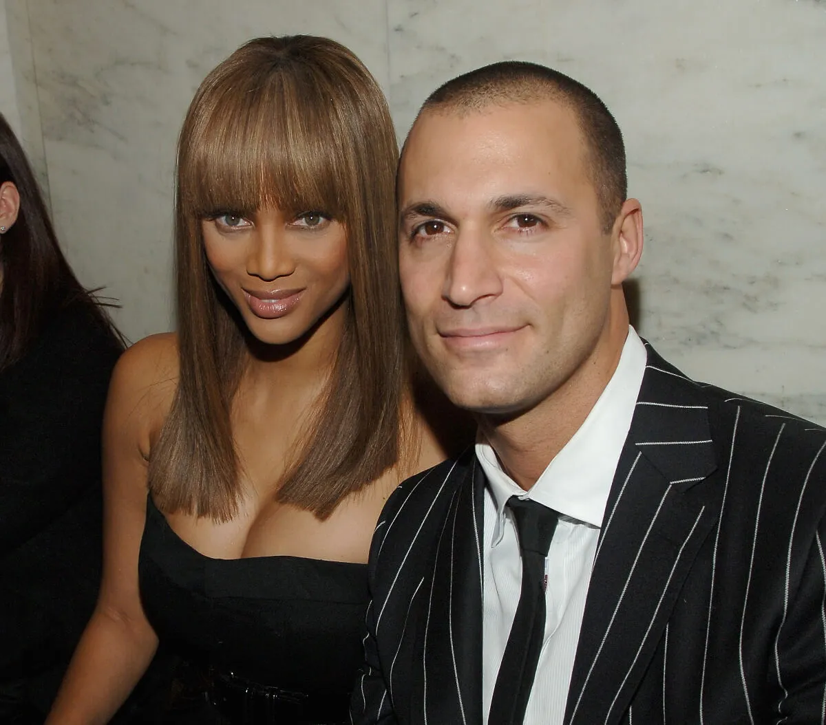 Tyra Banks wears black and sits with Nigel Barker, who wears a suit.