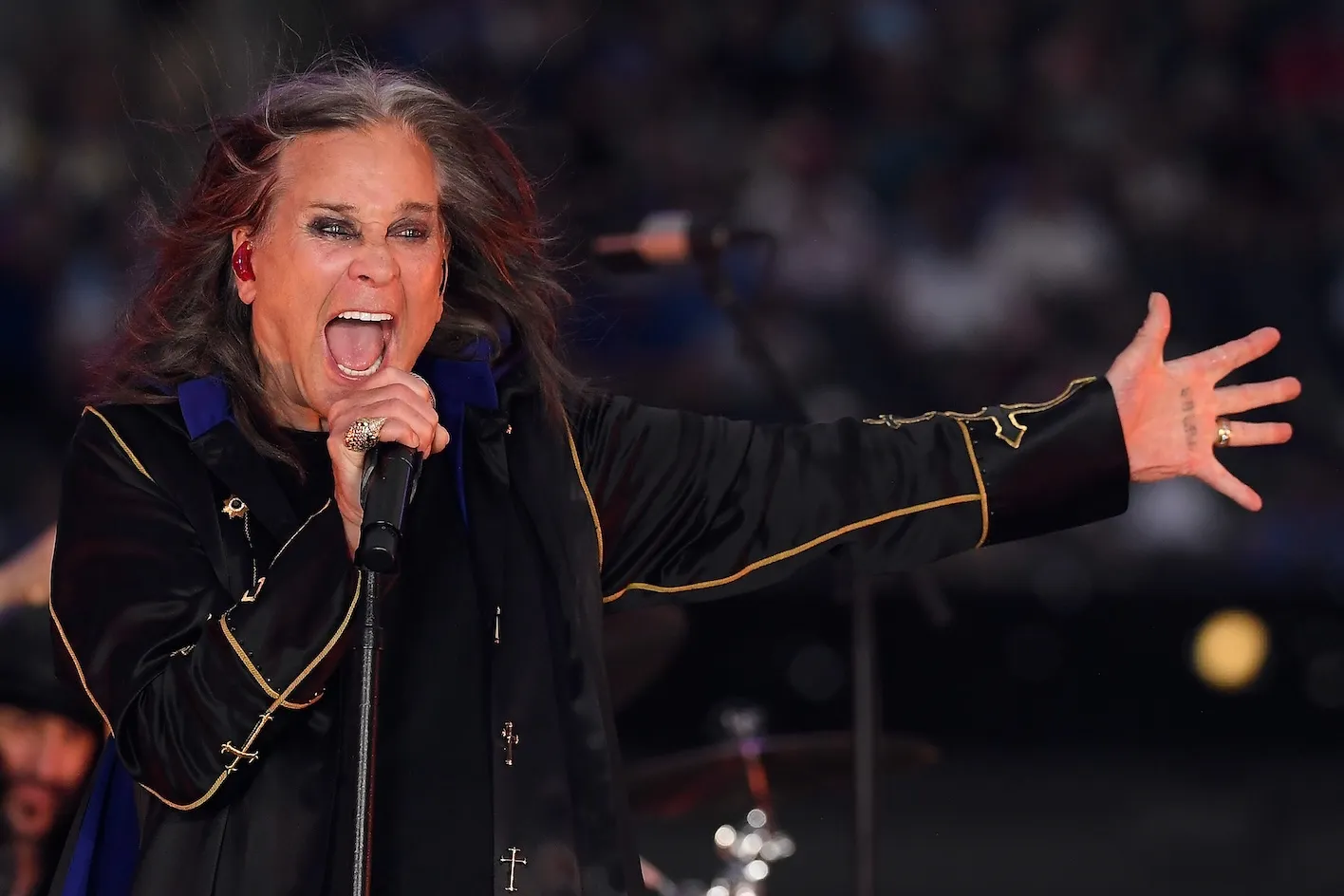 Ozzy Osbourne singing enthusiastically into a microphone with one arm arm and his fingers splayed.