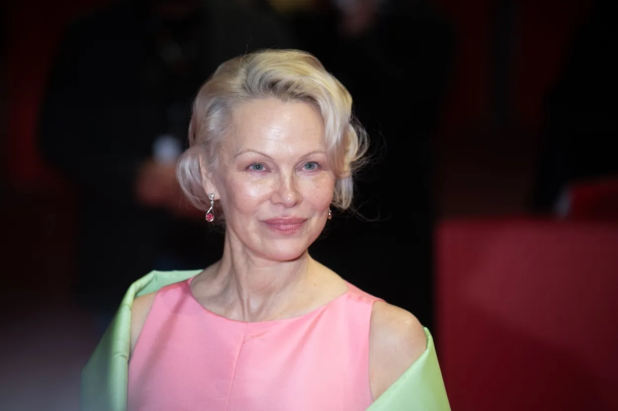 Pamela Anderson looking toward the camera in a pink dress with a green cover-up over her shoulders