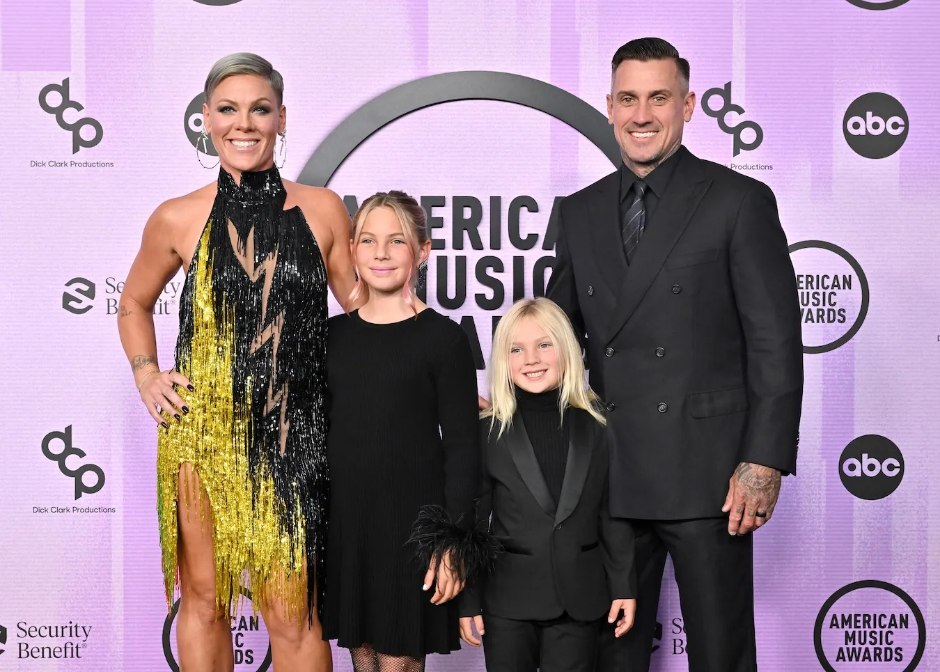 Pink, Carey Hart, and their 2 kids posing together in 2022. They are against a pink background and are dressed up.