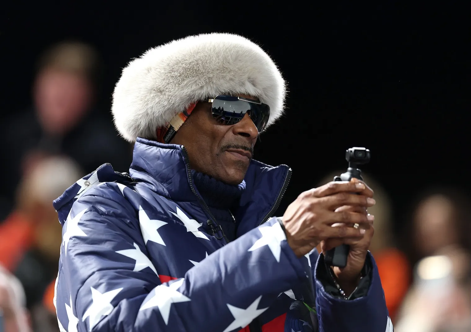 Snoop Dogg at the 2026 Winter Olympics. He's holding a camera up and wearing a USA-themed jacket and fur hat.
