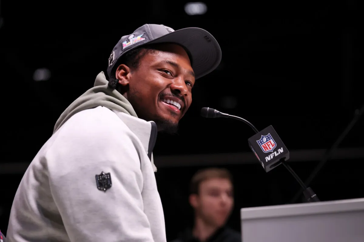 Stefon Diggs wears a white hoodie and a baseball hat. He sits in front of a microphone.
