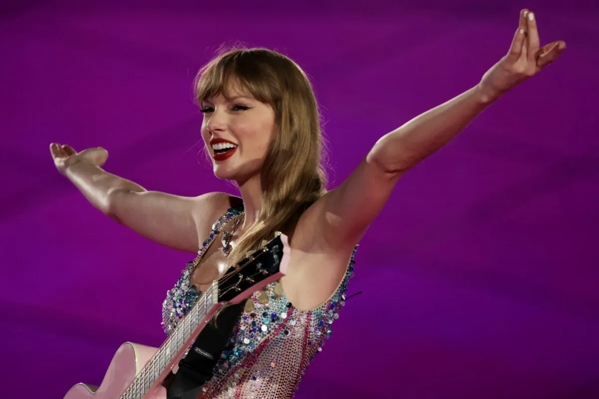 Taylor Swift wears a sparkly bodysuit and has an acoustic guitar hanging from her neck. She lifts her arms in the air.