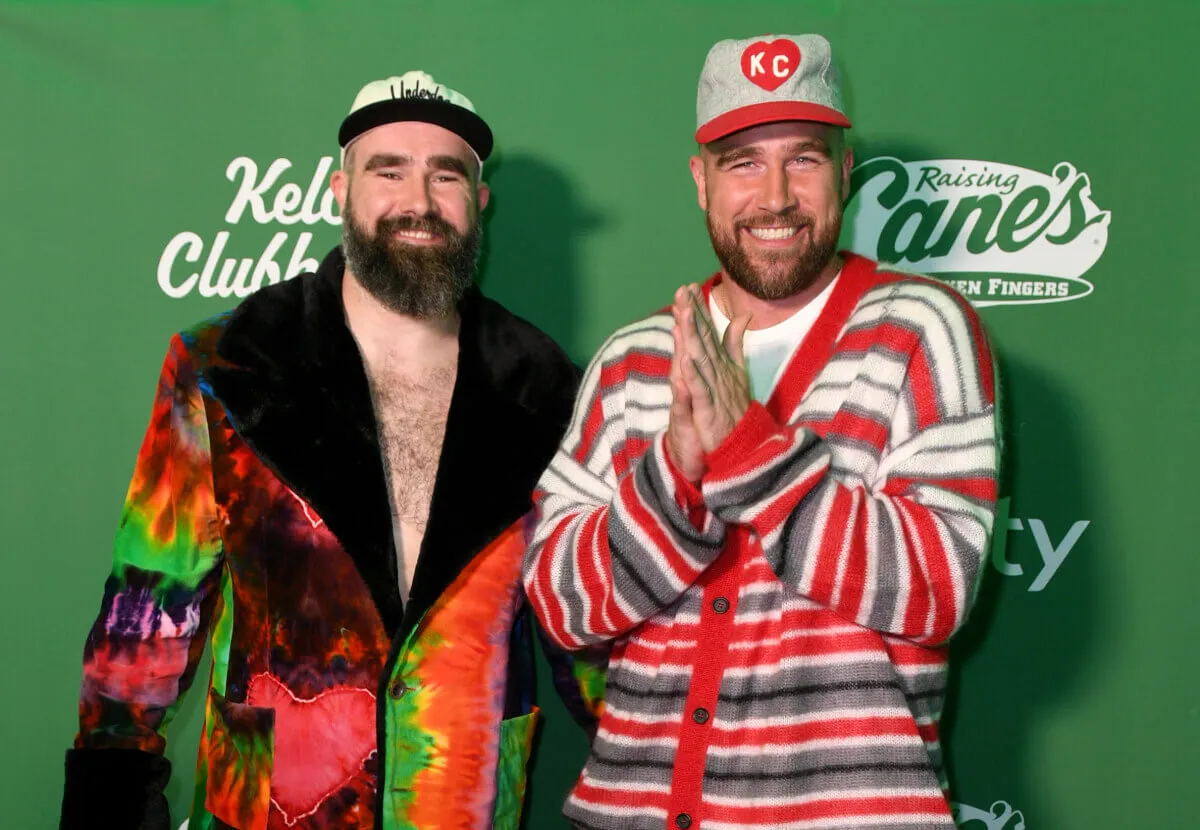 Jason Kelce wears a tie-dye jacket and stands with Travis Kelce, who wears a red striped sweater. They both wear baseball hats.