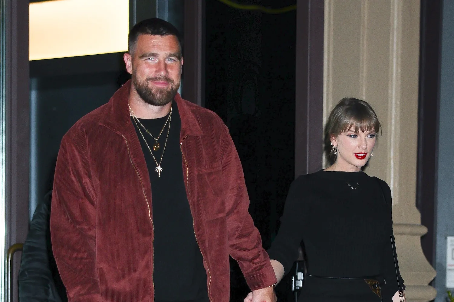Taylor Swift and Travis Kelce walking hand-in-hand in New York City in November 2025. Kelce is wearing a dark red jacket and Swift is in black.