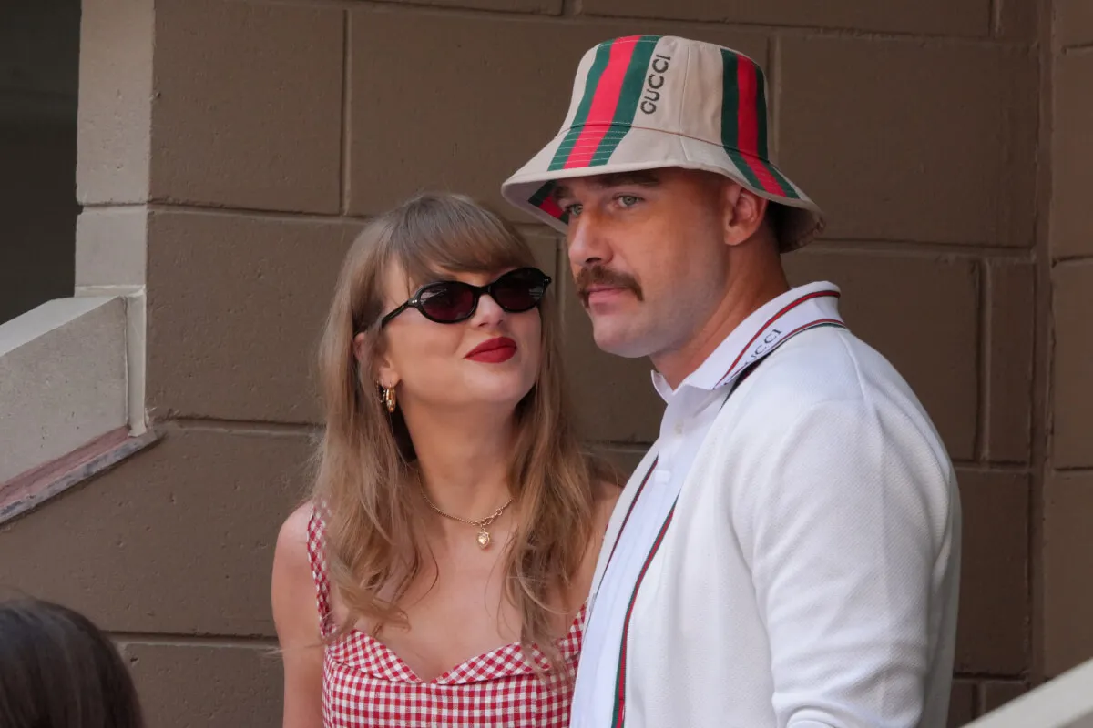 Taylor Swift wears a red gingham shirt and sunglasses. She looks up at Travis Kelce, who wears a white shirt and bucket hat.
