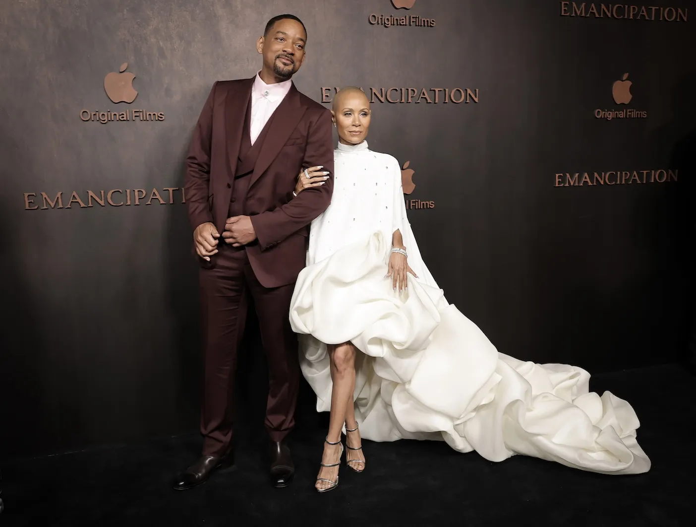 Will Smith in a dark maroon suit posing with Jada Pinkett Smith in a long-sleeve white dress
