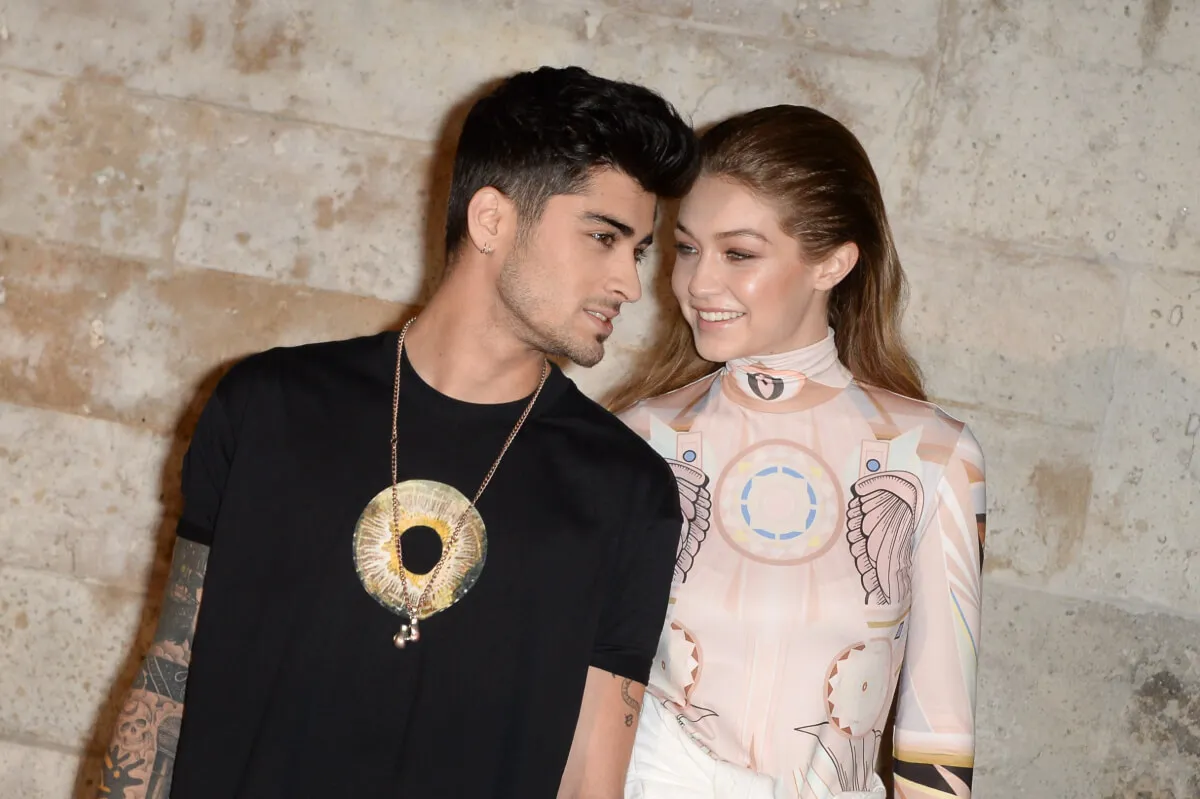 Zayn Malik wears a black shirt and stands with Gigi Hadid, who wears a white turtleneck.