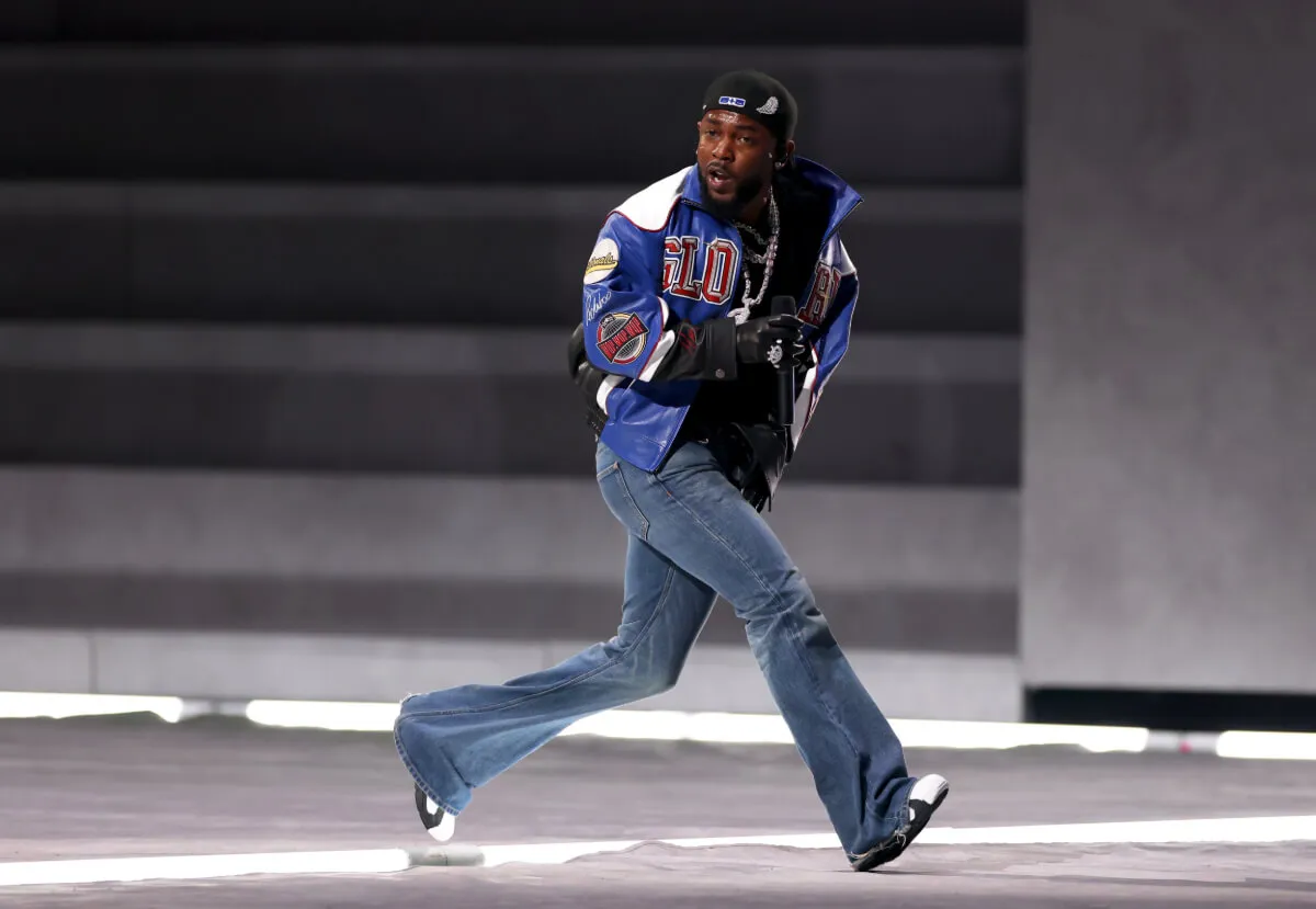 Kendrick Lamar wears jeans and a blue jacket. He runs across a stage during the Super Bowl.