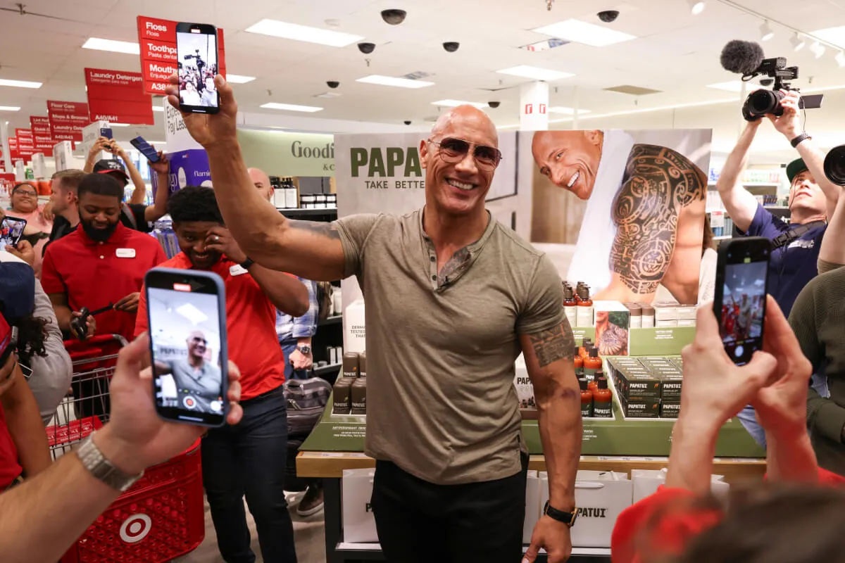 Dwayne Johnson stands in a Target and holds up a cell phone. There are people surrounding him.